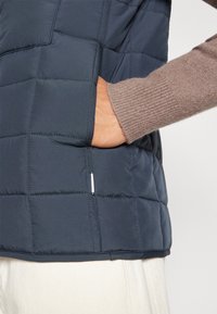 Vadderad marinblå väst med quiltat mönster. Har en sidoficka och en ärm i kolgrått stickat tyg, vilket framhäver kontrasterande texturer och färger.