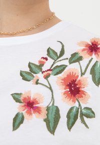 Bílé tričko s barevnou květinovou výšivkou v broskvových, červených a zelených tónech, s kulatým výstřihem a krátkými rukávy.