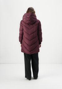 Burgunderfarget pufferjakke med sikk-sakk mønster, hette og ribbede mansjetter, kombinert med løse svarte bukser. Minimalistisk design på en ensfarget bakgrunn.