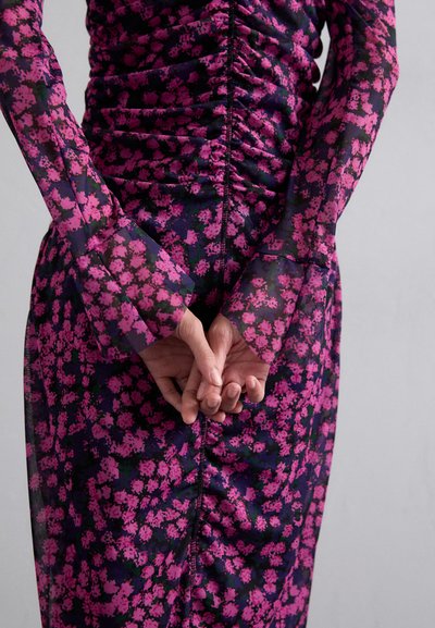 Robe �à manches longues à texture froncée, ornée d'un motif floral rose et violet sur un fond sombre. Mains jointes derrière le dos.