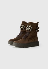 Stivaletti alla caviglia in suede marrone con suola platform, caratterizzati da accentuazioni decorative in gemme su una fascia, lacci frontali e dettagli con doppia fibbia.