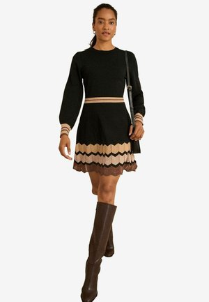 Schwarzes gestricktes Kleid mit langen Ärmeln, kontrastierendem beigen und braunen Zickzackmuster am Saum und einer taillierten Passform. Model trägt kniehohe Stiefel.