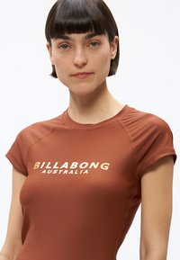 Fiatal nő rövid, sötét hajjal, rozsdabarna színű Billabong Australia pólót viselve, közvetlenül előre néz semleges arckifejezéssel.