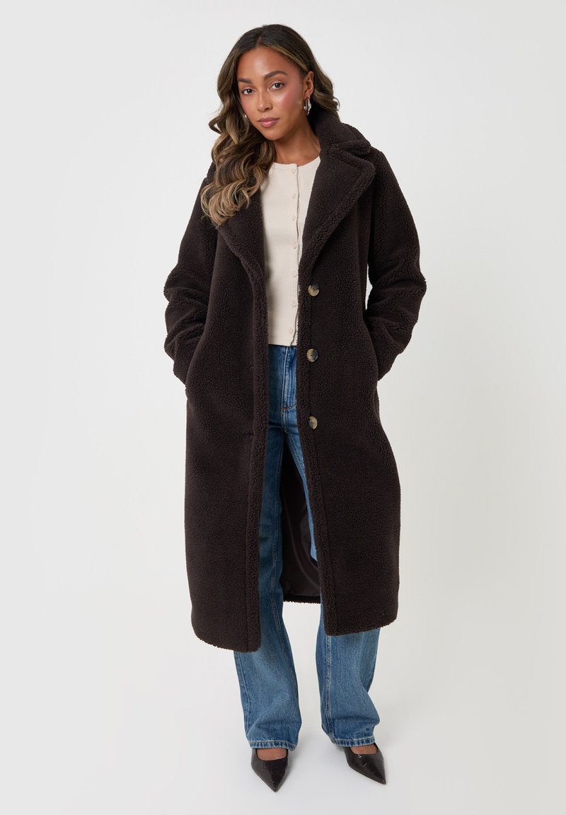 Woman with long wavy hair wearing a long dark fuzzy coat, light buttoned top, blue jeans, and black pointed shoes, standing with hands in pockets.
