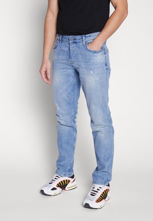 Man wearing light blue slim-fit jeans with small distressed patches and white sneakers with black and yellow details.