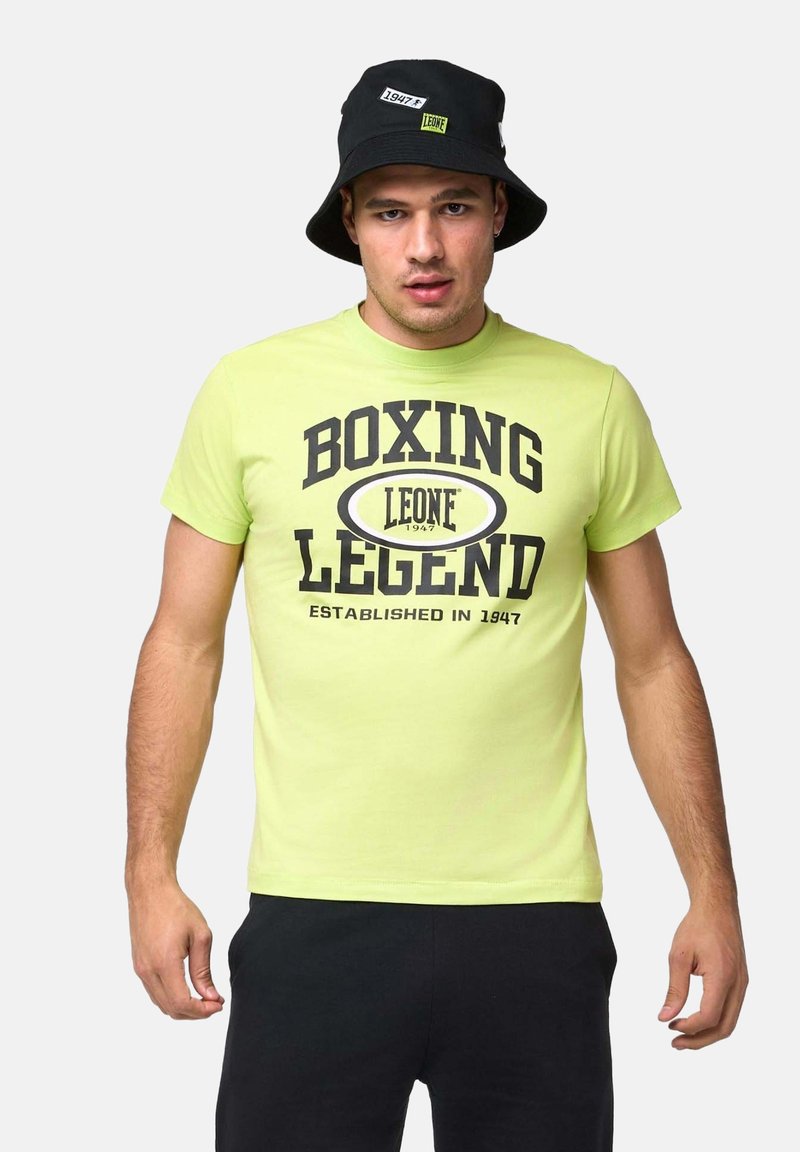 Homme portant un chapeau bob noir et un T-shirt vert citron avec le texte « Boxing Legend Leone Established in 1947 », debout devant un fond uni.