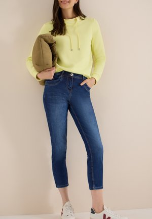 Kvinde iført korte blå jeans, gul sweatshirt, holder en olivengrøn dunjakke og hvide sneakers, stående mod en beige baggrund.