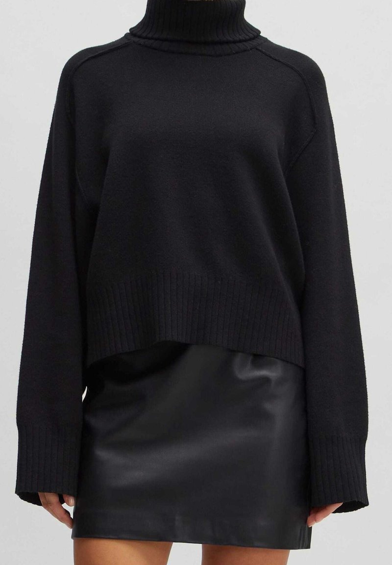Personne portant un pull à col roulé noir à manches longues et une mini-jupe en cuir noire sur un fond clair uni.