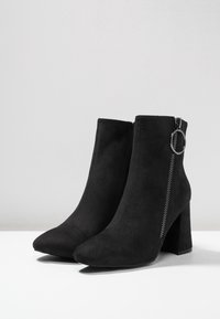 Botas de tobillo de ante negro con puntera afilada, cremallera lateral con detalle circular y un cómodo tacón cuadrado. Textura suave con un diseño elegante.