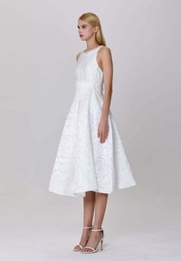 Femme aux longs cheveux blonds portant une robe midi sans manches à motifs floraux blancs et des sandales à talons hauts blanches, se tenant de profil.