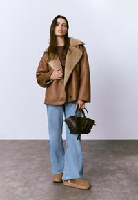 Mujer joven con una chaqueta de piel de oveja marrón, camisa a rayas, pantalones vaqueros de pierna ancha azul claro, zapatos de lona beige, sosteniendo un bolso de mano marrón oscuro.