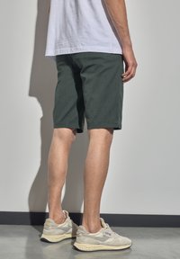 Pantalones cortos verdes oscuros hechos de tela texturizada, que terminan justo por encima de la rodilla, combinados con zapatillas de color claro. El fondo incluye una pared clara.