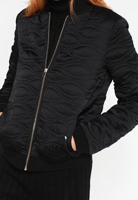 Czarna pikowana kurtka bomberka z falistymi wytłoczonymi wzorami, złotym zapięciem na zamek i prążkowanymi mankietami oraz dołem. Miękka, fakturowana tkanina.