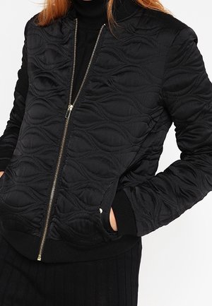 Chaqueta bomber acolchada negra con patrones ondulados en relieve, cierre de cremallera dorada y puños y dobladillo de canalé. Tejido suave y texturizado.