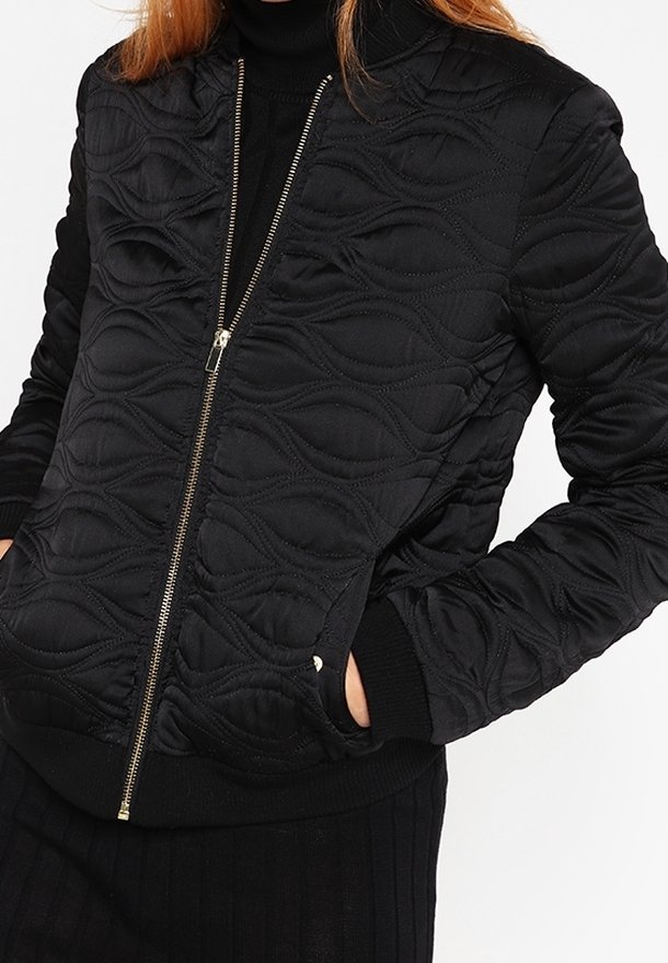 Czarna pikowana kurtka bomberka z falistymi wytłoczonymi wzorami, złotym zapięciem na zamek i prążkowanymi mankietami oraz dołem. Miękka, fakturowana tkanina.