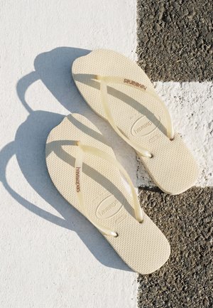 Chanclas Havaianas beige colocadas sobre una superficie texturizada blanca y negra con la luz del sol proyectando sombras.
