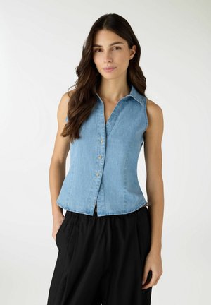 Donna con lunghi capelli castani indossa una camicia di jeans senza maniche azzurra chiusa con bottoni e pantaloni neri larghi, in piedi davanti a uno sfondo neutro.