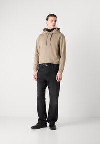 Felpa beige con cappuccio e lacci, jeans neri in denim e scarpe nere. Texture morbida, design casual e vestibilità rilassata. Sfondo semplice.