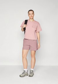Camicia rosa chiaro a maniche corte con tasca frontale, abbinata a pantaloni corti coordinati. Indossata con sneakers grigie e uno zaino nero.