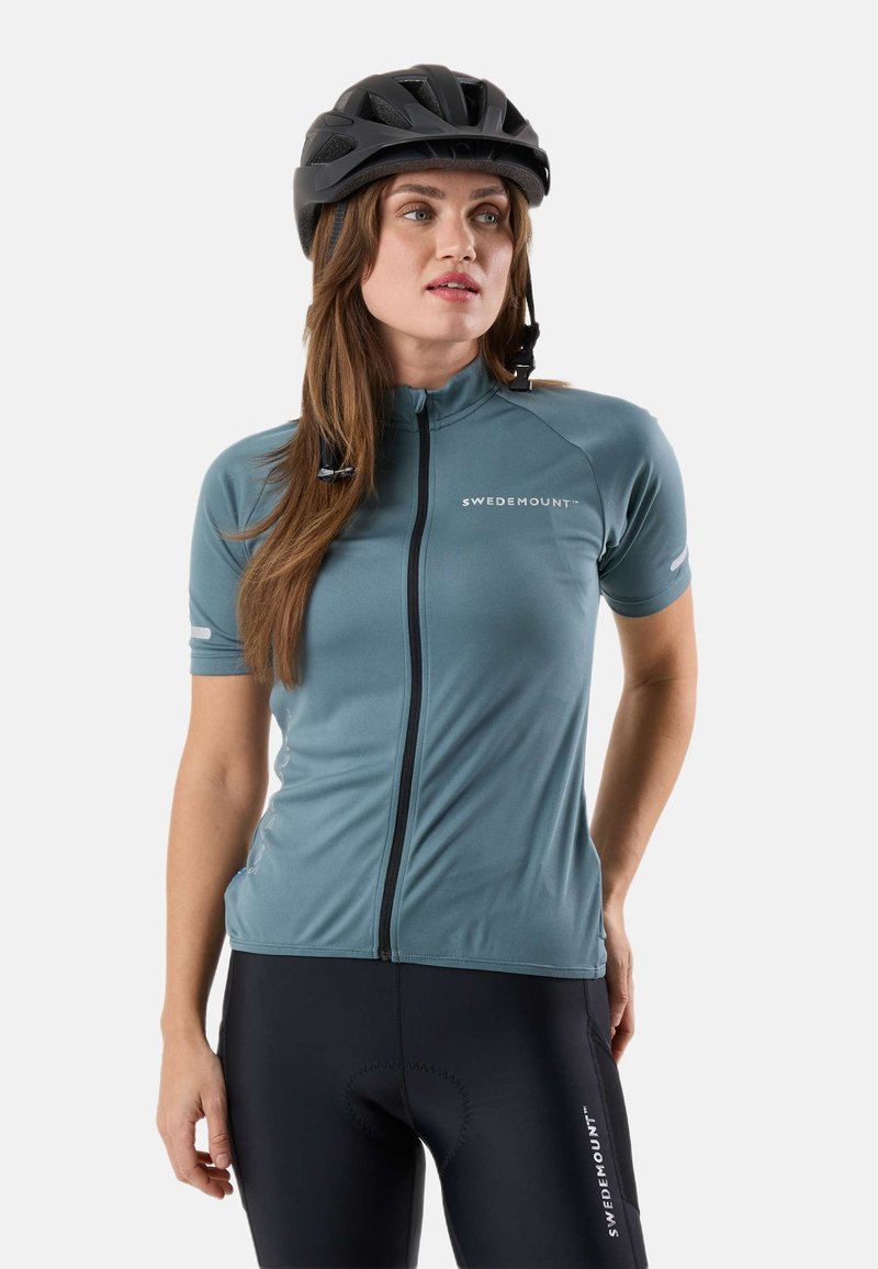 Femme portant un maillot de cyclisme bleu à manches courtes et un casque noir, debout les mains derrière le dos devant un fond uni.