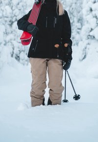 Personne en veste noire et pantalon beige tient des skis rouges et des bâtons de ski, debout dans une neige profonde avec des arbres enneigés en arrière-plan.