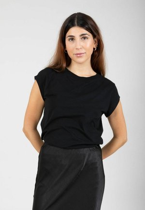 Vrouw met lang bruin haar, gekleed in een zwart shirt met korte mouwen en een zwarte rok, staand met haar handen achter haar rug tegen een effen achtergrond.