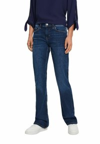 Jean en denim bleu foncé avec une coupe droite, un design classique à cinq poches et des coutures contrastées, porté avec des baskets blanches.