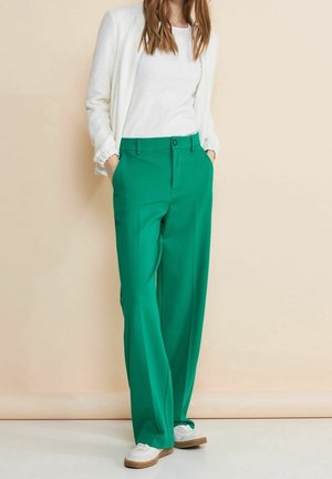 Vrouw die een witte top, een gestructureerde witte jas, felgroene broek met wijde pijpen en witte sneakers draagt, staand met haar handen in haar zakken.