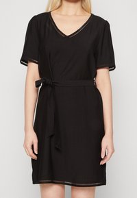 Nainen, jolla on lyhythihainen musta mekko, jossa on V-kaula-aukko, vyötärösolki ja koristeelliset leikkuureunat hihoissa, kaula-aukossa ja helman reunassa.