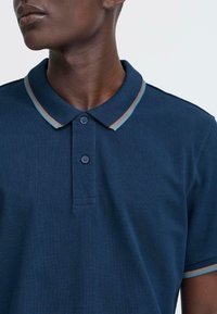 Moški nosi mornarsko moder polo majico z svetlo modrimi in rjavimi črtami na ovratniku in robovih rokavov, gumbi zaprti pri vratu.