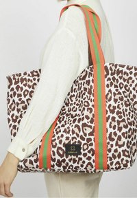 Torebka tote w leopardzie wzorze w odcieniach brązu i kremu, z pomarańczowo-zielonymi paskami. Na boku widoczna jest czarna metka z logo.