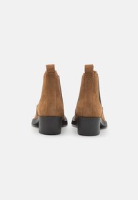 Botines de ante marrón con un tacón negro liso, que cuentan con una sutil etiqueta de logo en la parte trasera y un diseño elegante sin cordones.