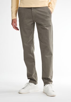 Mann trägt graue Freizeithose mit teilweise in den Taschen steckenden Händen und weißen Turnschuhen, steht vor einem schlichten Hintergrund.