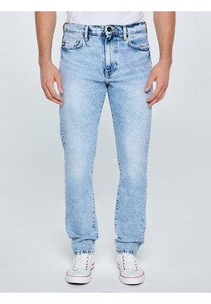 Vyras dėvi šviesiai mėlynas tiesaus kirpimo džinsus ir baltus sportbačius, stovi prie paprasto fono, rankos laisvai nuleistos šonuose.