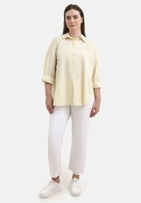 Camicia a righe gialla e lavanda con bottoni frontali e maniche arrotolate, abbinata a pantaloni bianchi e sneakers bianche.