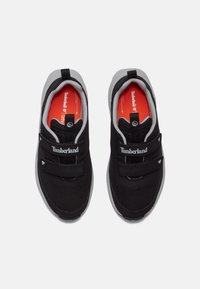 Baskets noires avec une tige en mesh et des accents gris, dotées de sangles Velcro ajustables, d'une semelle intérieure rouge et du logo Timberland sur le côté.