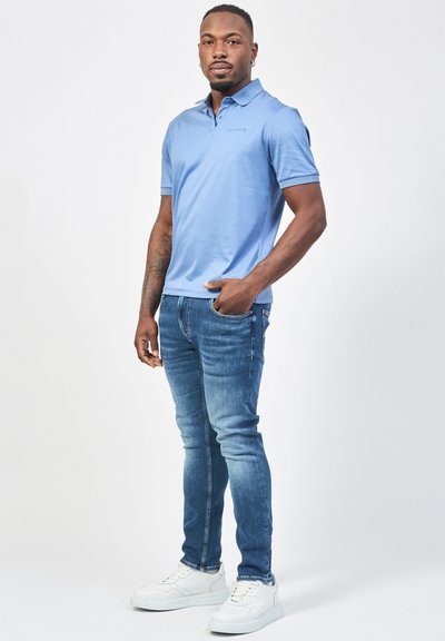 Polo shirt azzurro chiaro con maniche corte e colletto, abbinato a jeans blu slim-fit e sneaker bianche. Presenta un branding minimal sulla maglietta.