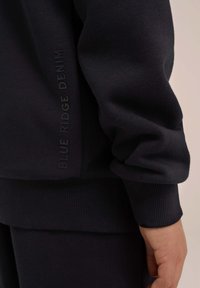 Temno modra trenerka z rebrastimi manšetami in robom. "BLUE RIDGE DENIM" natisnjeno na strani. Mehka tekstura in sproščen kroj.