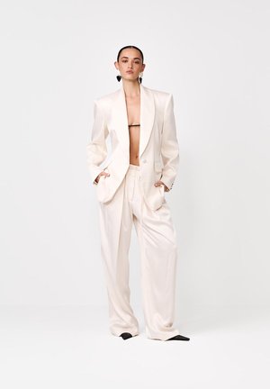 Femeie care poartă un costum oversize de culoare crem deschis, cu pantaloni largi, fără cămașă, cu un bandeau negru minimal, mâinile în buzunare și pantofi negri cu vârf ascuțit.