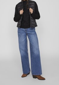 Svart stickad tröja, grå vadderad väst, vida blå jeans och bruna klackboots. Enkel design med en avslappnad passform.