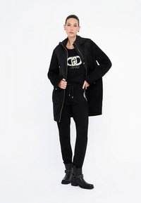 Veste matelassée noire avec capuche, superposée à un T-shirt noir avec un logo. Joggeurs noirs ornés et bottines à la cheville.