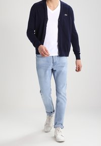 Mörkblå knäppbar cardigan över en vit V-ringad t-shirt, matchad med ljusblå jeans och vita sneakers. Har en liten logotyp på cardiganen.