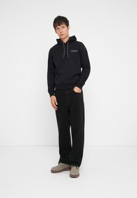Young man standing in casual black hoodie, black pants, and beige boots against white background with one hand in pocket.