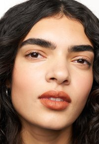 Gros plan sur un visage avec des cheveux ondulés et foncés. Caractéristiques des sourcils fournis et bien définis, des lèvres mates aux tons chauds et une texture de peau lisse.