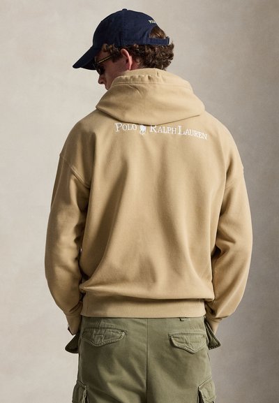 Άντρας με μπεζ φούτερ Polo Ralph Lauren, χακί cargo παντελόνι, μαύρο καπέλο και γυαλιά ηλίου, γυρισμένος με την πλάτη σε απλό φόντο.