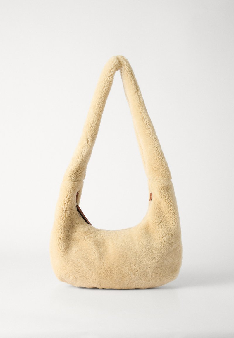 Beige särgmed kattega õlaline kott, millel on kumer käepide ja ümar kuju. Tekstuur on pehme ja plüüsine, sisaldades minimaalset metallist aksessuaari.