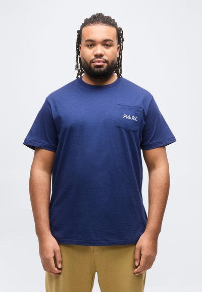 Άνδρας με πλεξούδες στα μαλλιά και γενειάδα, που φοράει μπλε μπλουζάκι Polo RL και μπεζ παντελόνι, στέκεται μπροστά σε λευκό απλό φόντο.