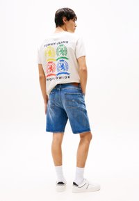 Grå T-skjorte med flerfargede løve-logoer på baksiden, kombinert med blå denimshorts. Hvite joggesko og synlige hvite sokker.