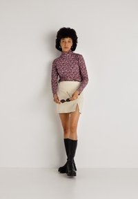 Blusa de cuello alto de manga larga, morada con estampado floral, combinada con una minifalda beige con una abertura lateral, complementada con botas negras hasta la rodilla.