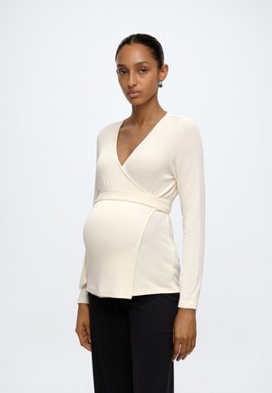 Femme enceinte portant un top cache-cœur couleur crème et un pantalon noir, debout devant un fond clair uni.